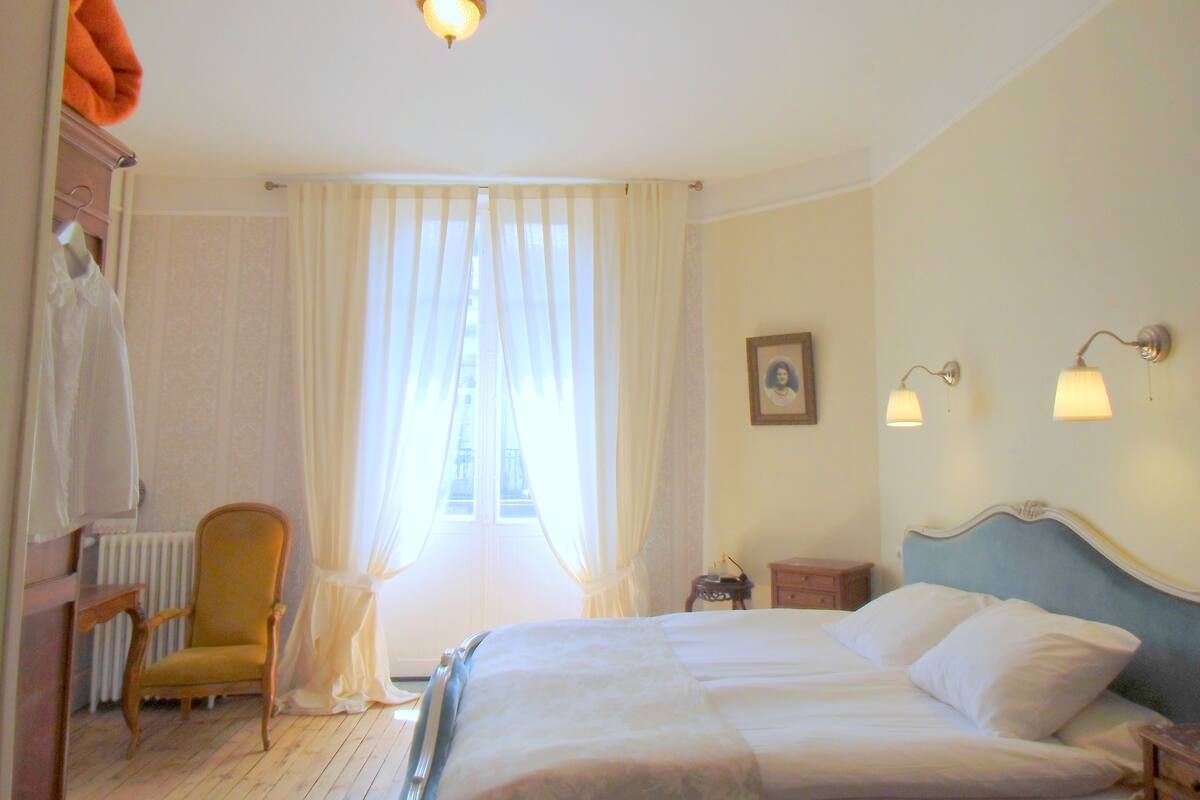 Habitación de hotel con cama, una silla, una mesita de noche, un radiador y una ventana con cortinas.