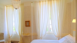 Quarto com cama grande, cortinas brancas, um candelabro e uma pequena mesa de cabeceira.