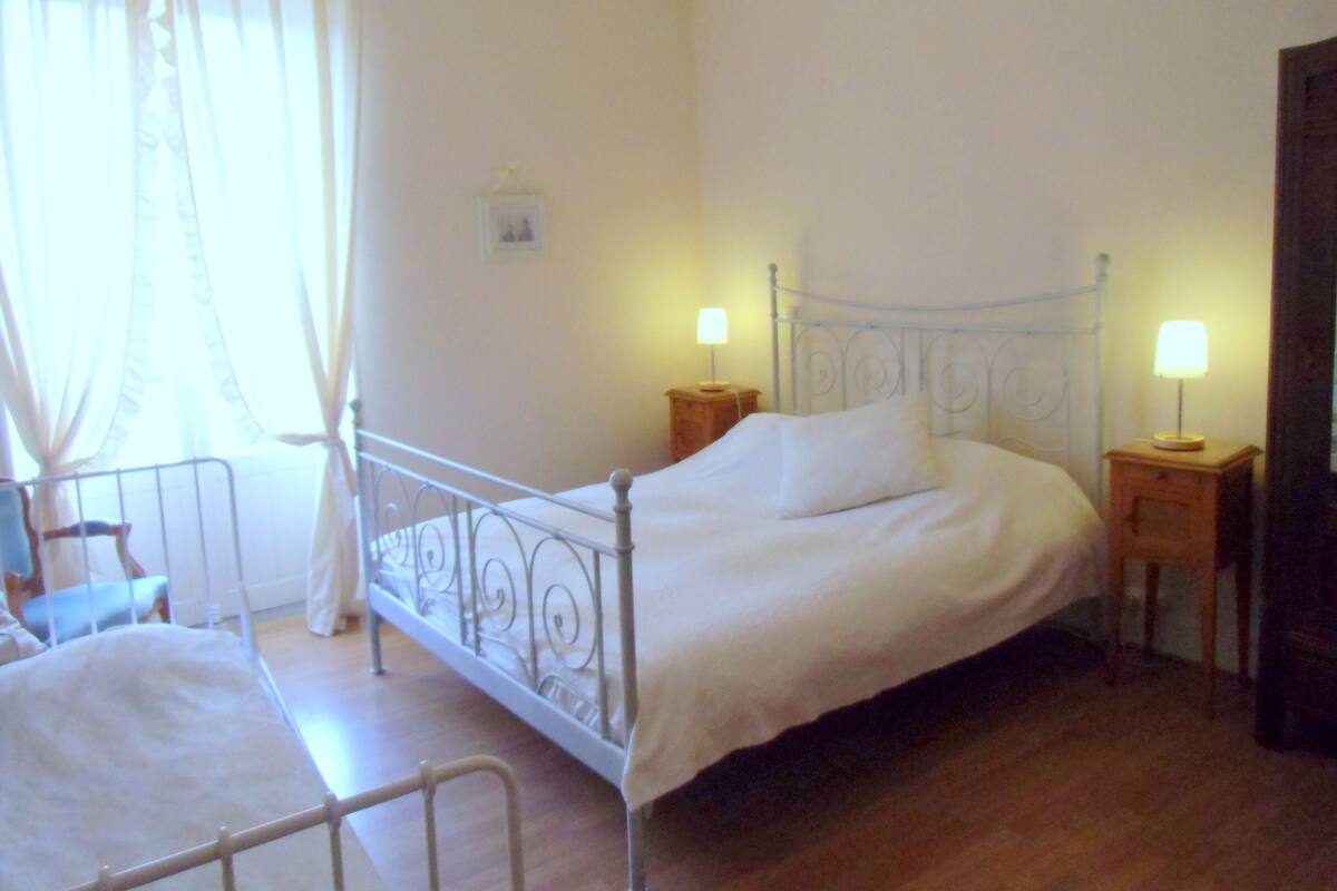 Una cama individual con estructura de metal, dos mesitas de noche con lámparas, piso de madera y una ventana con cortinas traslúcidas.