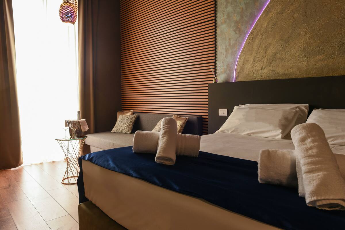 A hotel room with a large bed, a bedside table with a lamp, and a view of the outside through the window.