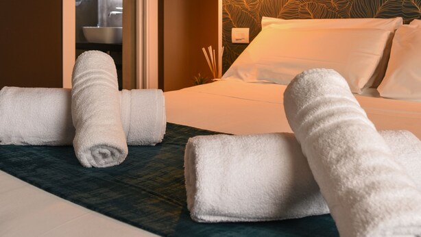 Habitación de hotel con cama, toallas blancas enrolladas, una puerta que da a un baño y una pared con diseño de hojas.