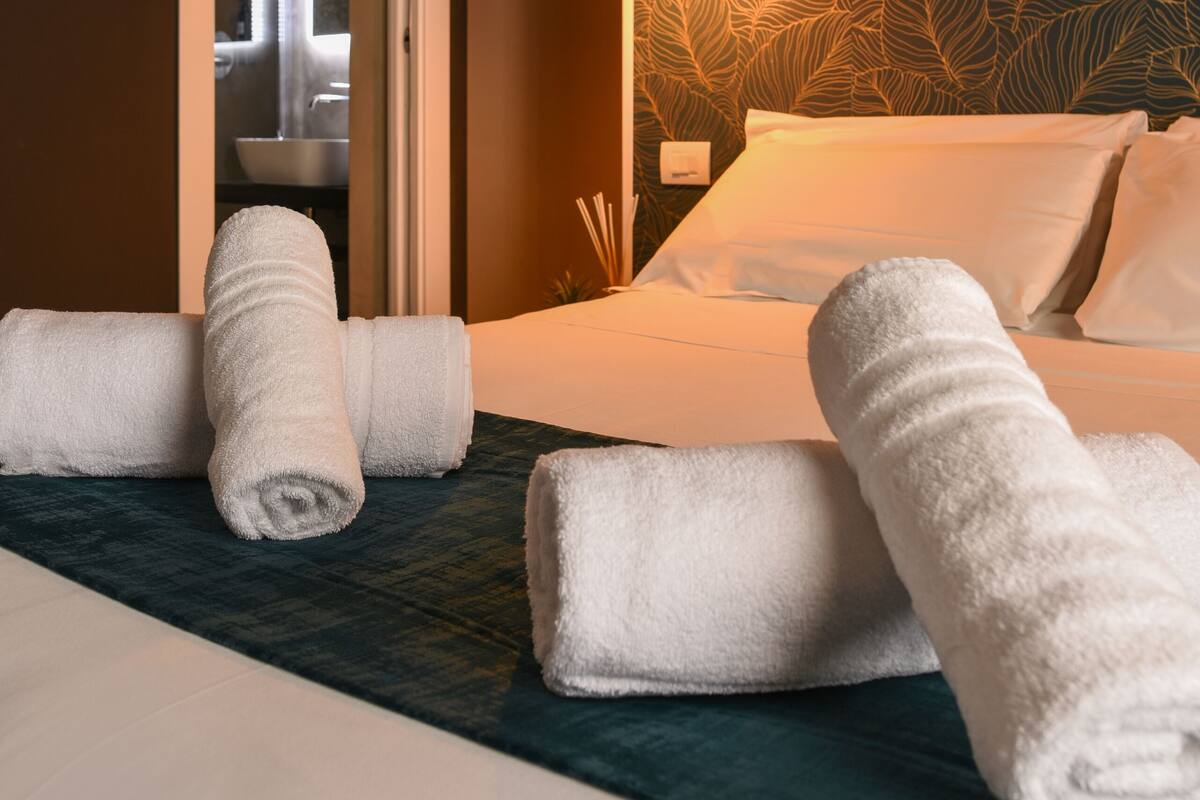 A hotel room with a bed, rolled white towels, a door leading to a bathroom, and a wall with a leafy pattern.