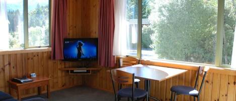 A cozy cabin room with a television, wooden walls, a table with chairs, and large windows offering a view of trees.