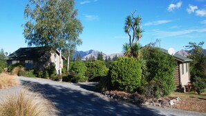 Une route de campagne menant à une maison entourée d’une végétation luxuriante, avec des montagnes en arrière-plan.