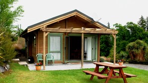 Une cabane en bois avec un porche couvert, deux chaises vertes, une table de pique-nique et des plantes en pot.