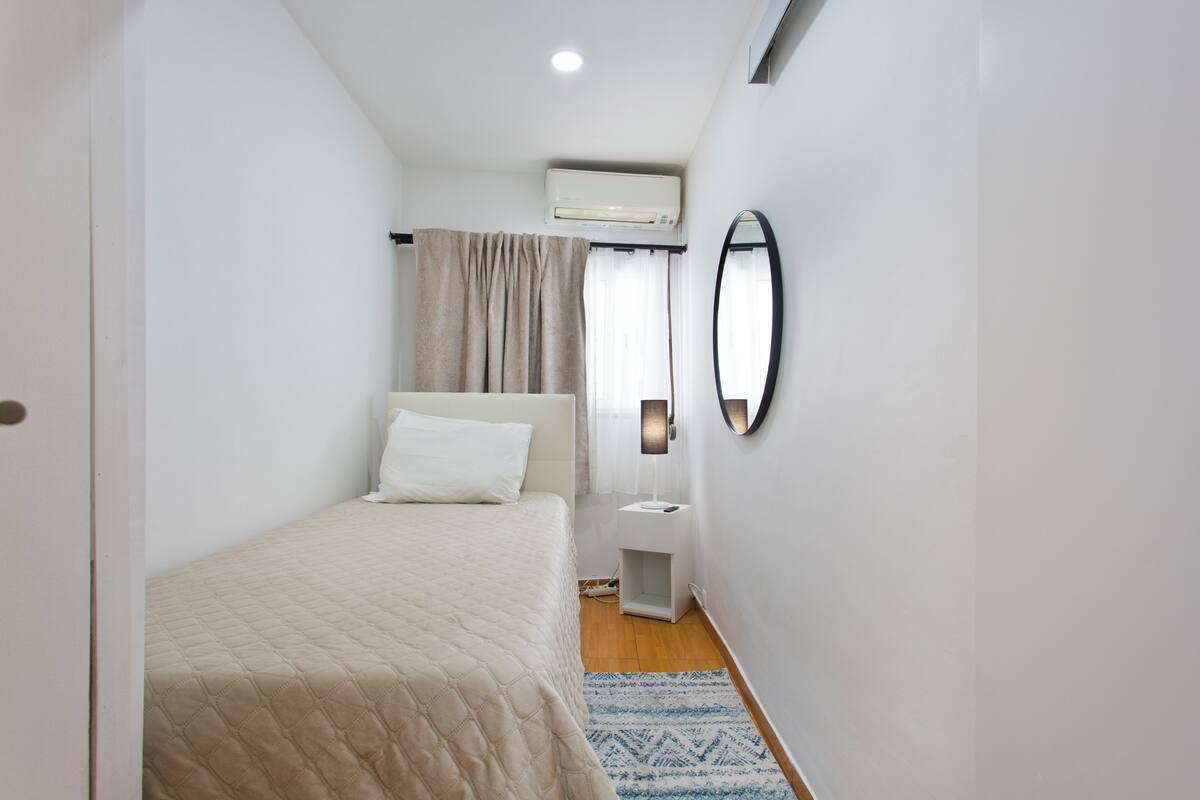 A bedroom with a bed, bedside table, wall-mounted mirror, and an air conditioning unit.
