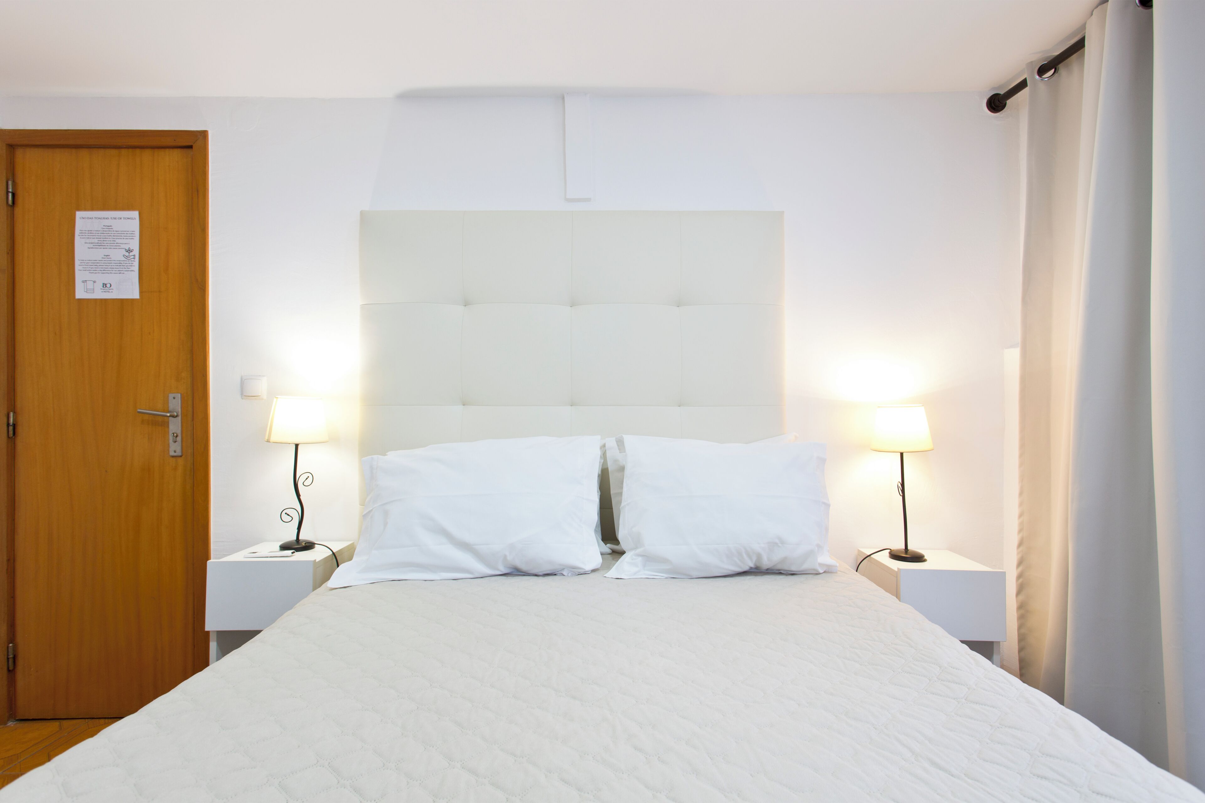 A hotel room with a bed, two bedside tables with lamps, a wooden door, and a white headboard.