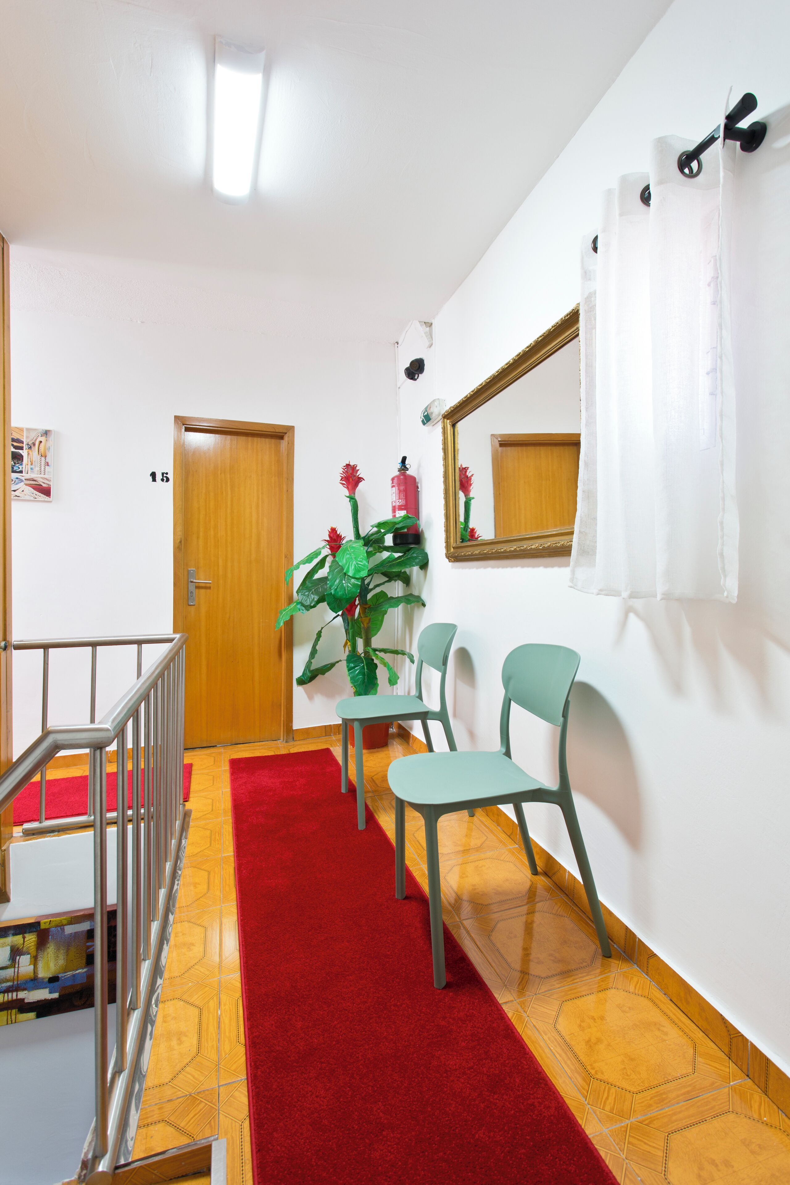 Un couloir avec un tapis rouge, une porte en bois, des chaises vertes, une table, une plante en pot et un miroir au mur.