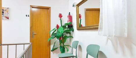 Un couloir avec un tapis rouge, une porte en bois, des chaises vertes, une table, une plante en pot et un miroir au mur.