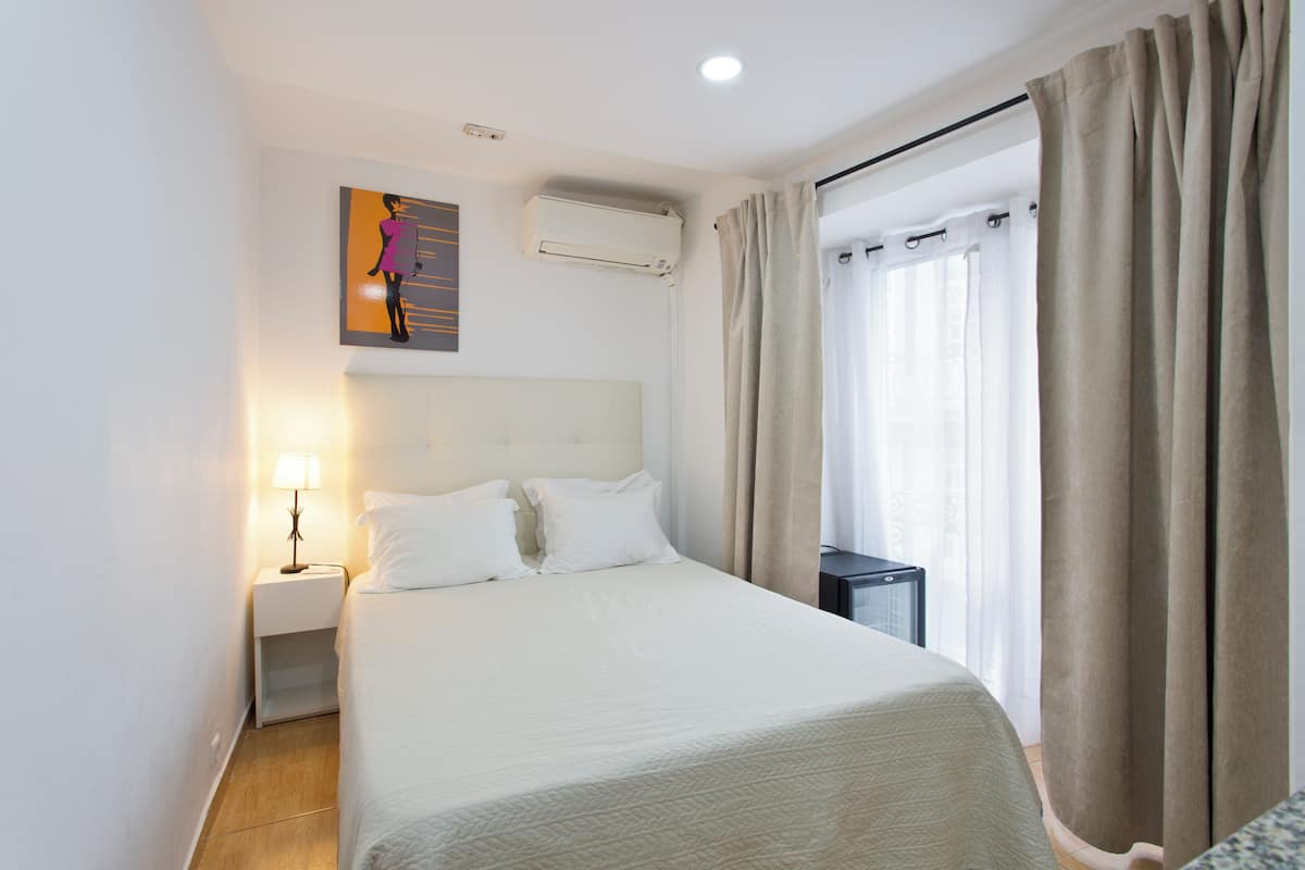 A hotel room with a bed, a nightstand with a lamp, an air conditioning unit, and a window with curtains.