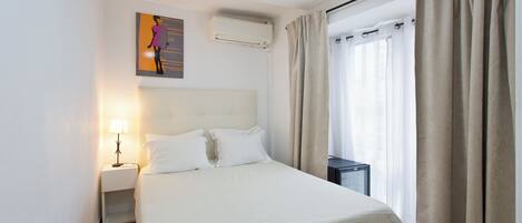 A hotel room with a bed, a nightstand with a lamp, an air conditioning unit, and a window with curtains.