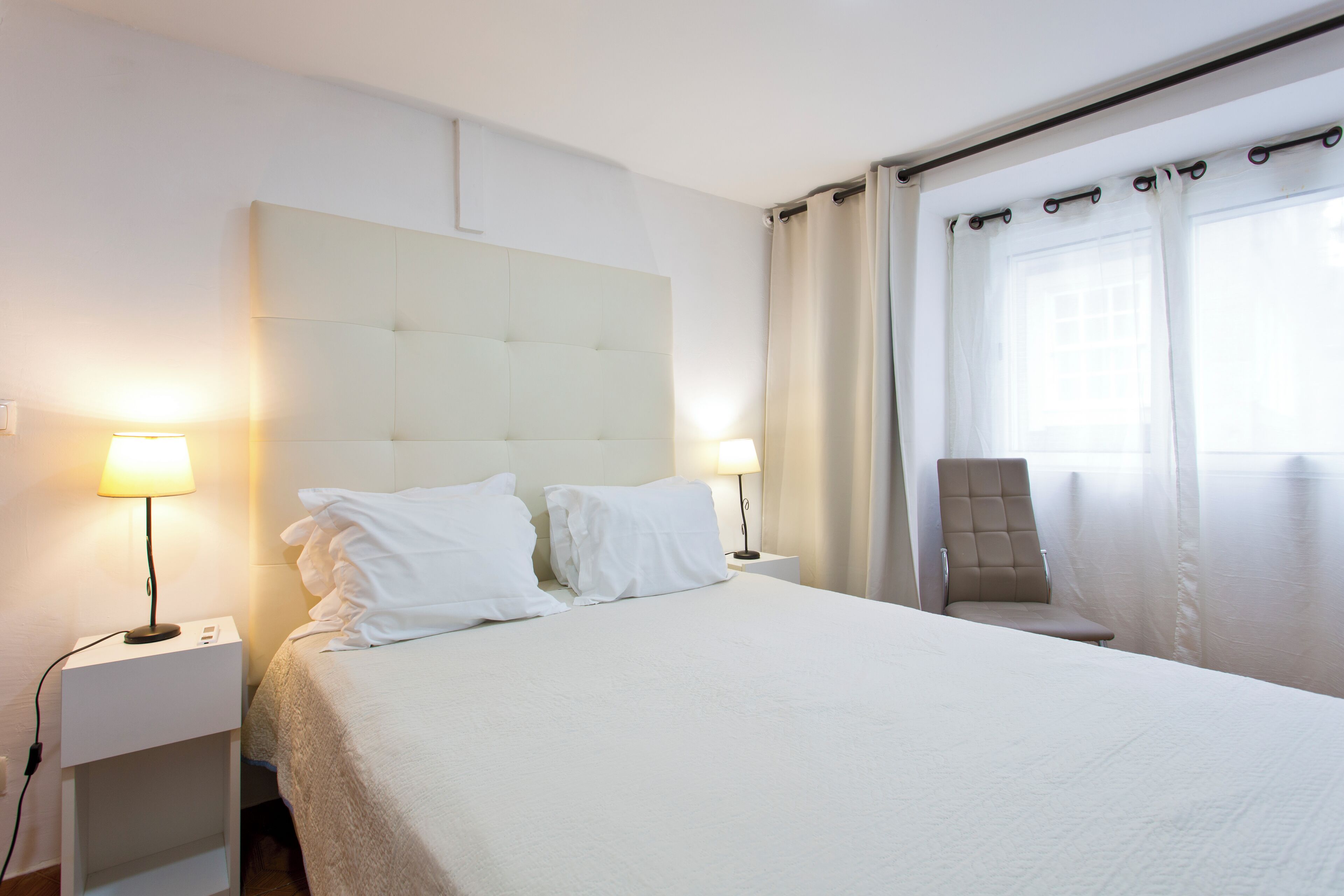 Una cama bien hecha con ropa de cama blanca, dos lámparas de noche, una silla y una ventana con cortinas.