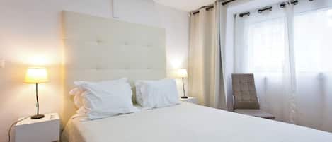 Una cama bien hecha con ropa de cama blanca, dos lámparas de noche, una silla y una ventana con cortinas.