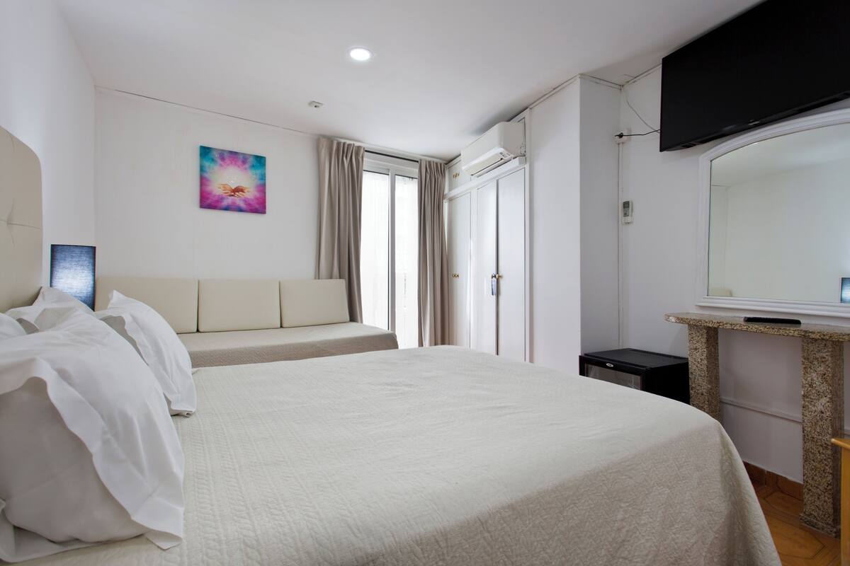 A hotel room with a large bed, a television mounted on the wall, a window with curtains, and a small table with a mirror.