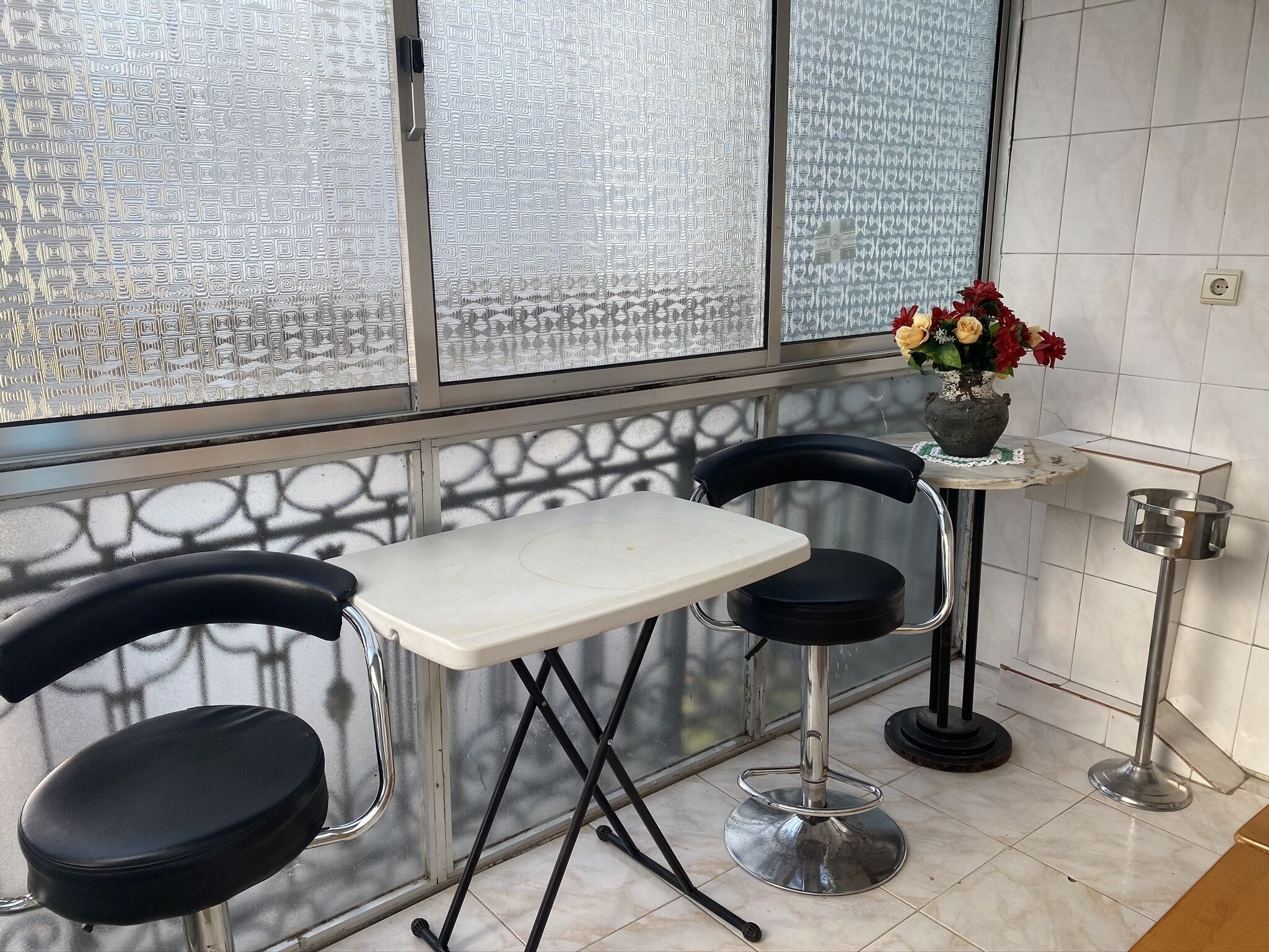 Eine kleine, moderne Küche mit einem weißen Tisch und zwei schwarzen Stühlen, einem kleinen runden Tisch mit Blumen in einer Vase und einem Metallgestell mit einem Topf.