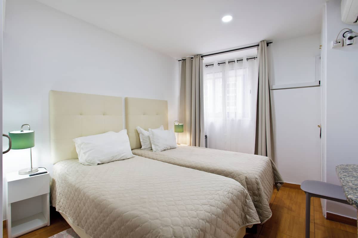 A hotel room with two beds, a bedside table with a lamp, a chair, and a window with curtains.