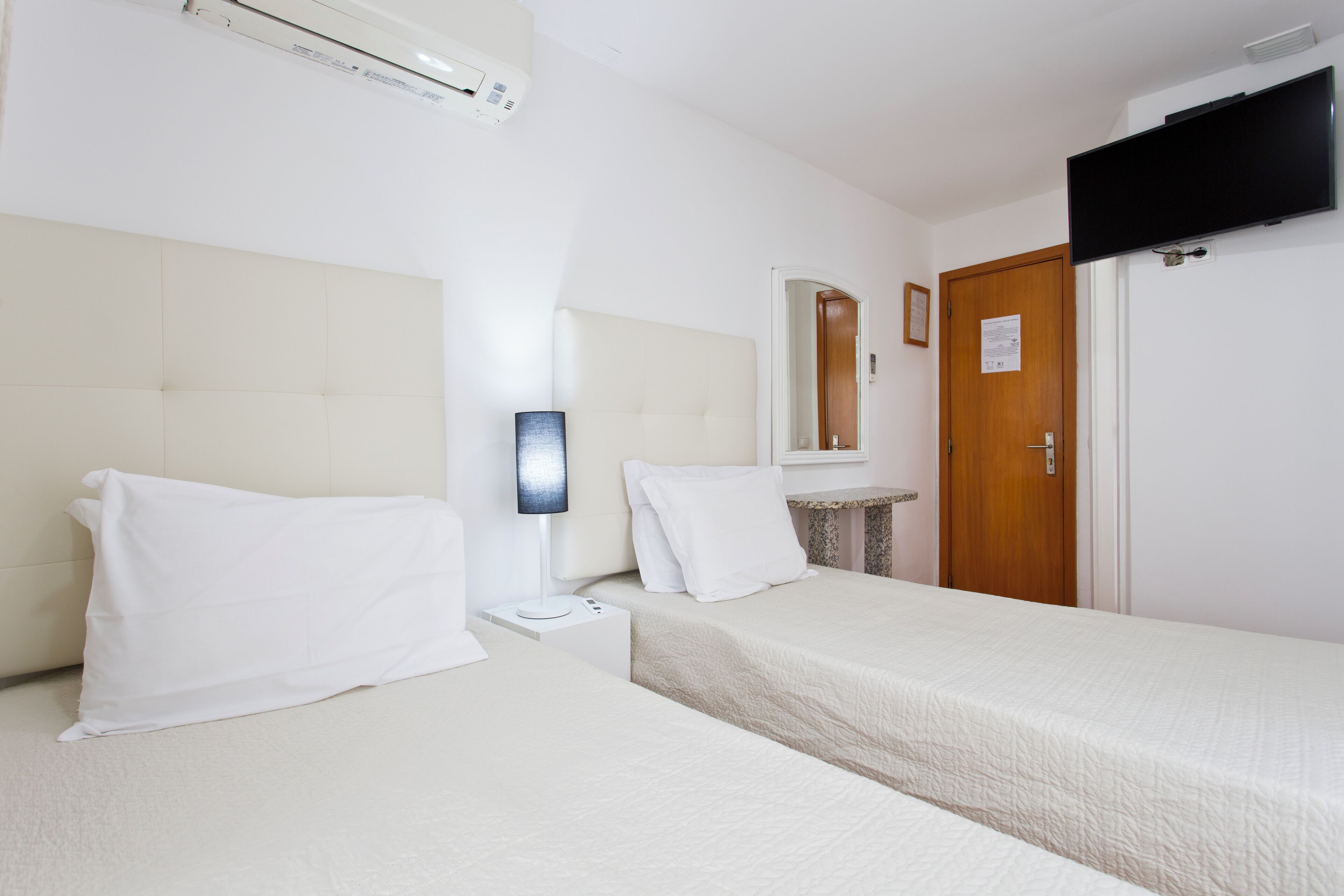 A hotel room with two beds, a flat-screen TV mounted on the wall, a bedside table with a lamp, and a wooden door.