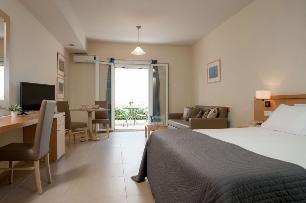 A hotel room with a bed, desk, chair, and a small table with a chair. There is a television on the desk, a window with curtains, and a view of the outside.