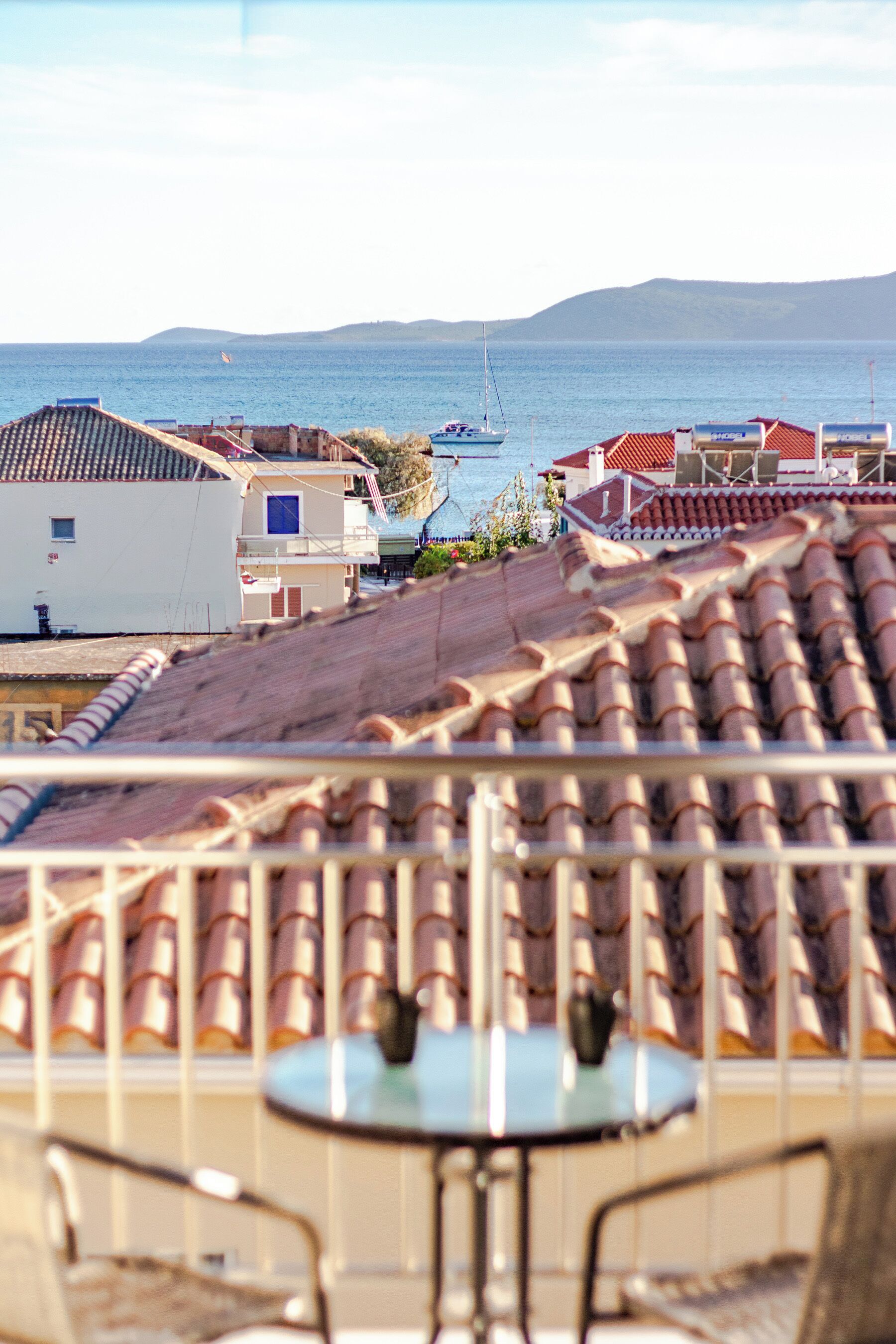 A terrace with a table and chairs, overlooking a coastal area with buildings and a sailboat in the distance.