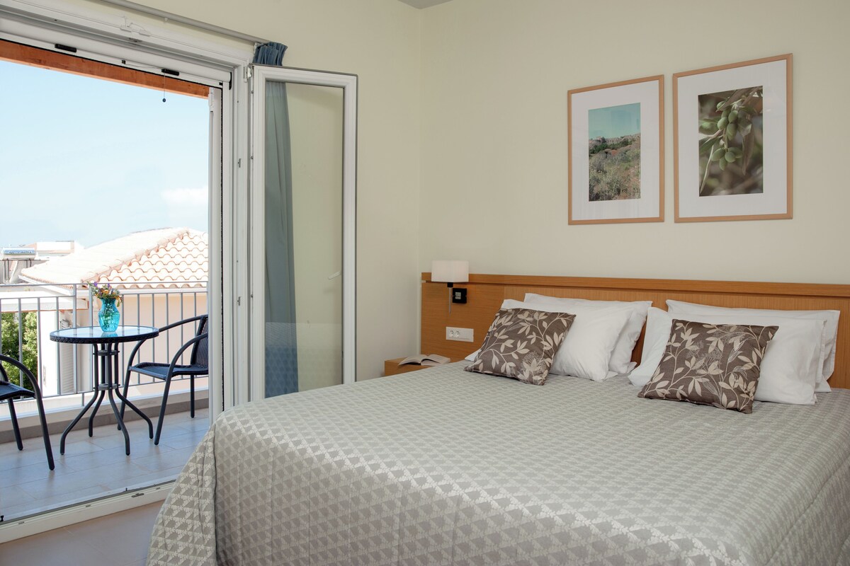A hotel room with a bed, two framed pictures on the wall, a balcony with a table and chairs, and a view of the outside.