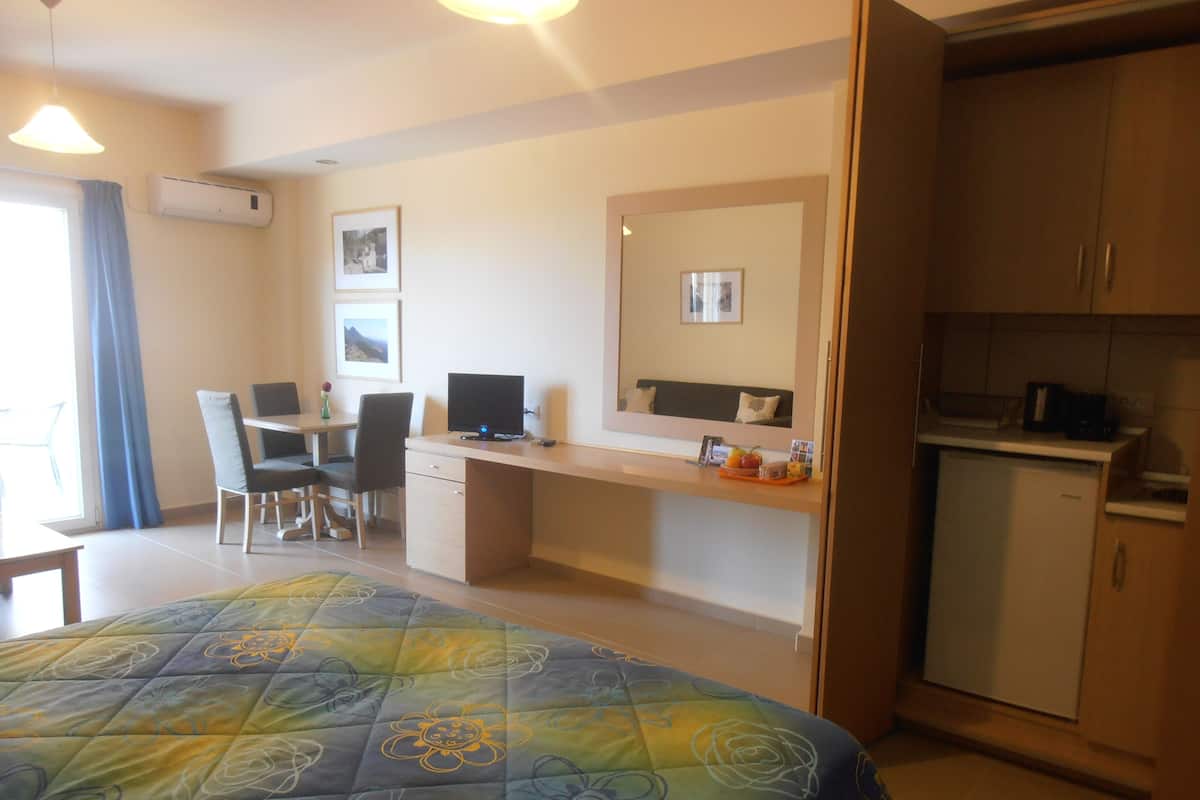 A hotel room with a bed, a desk with a computer, a chair, a small kitchenette, and a window with curtains.