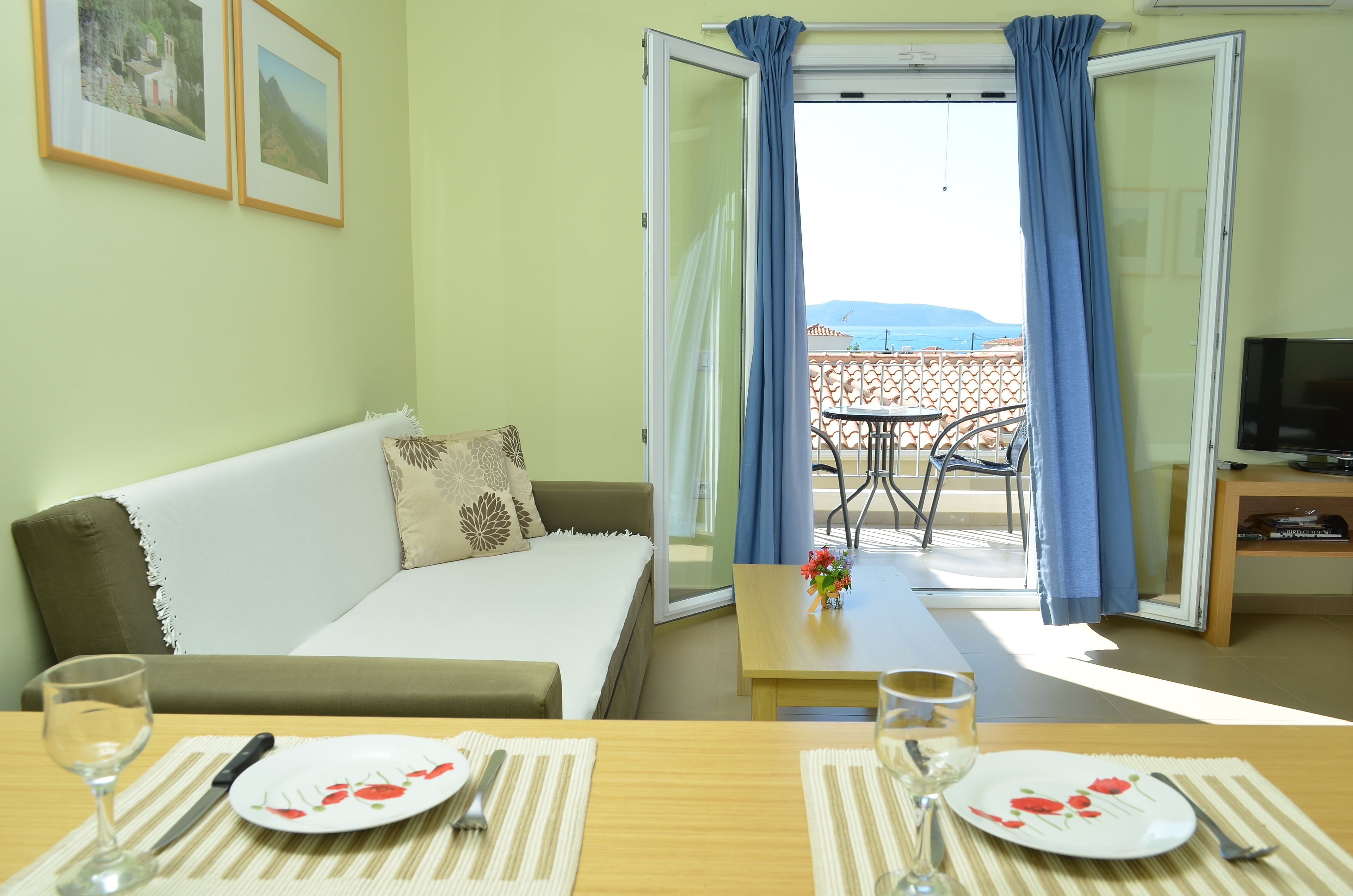 A room with a bed, a dining table set with plates and cutlery, a TV stand, and a balcony with a table and chairs.