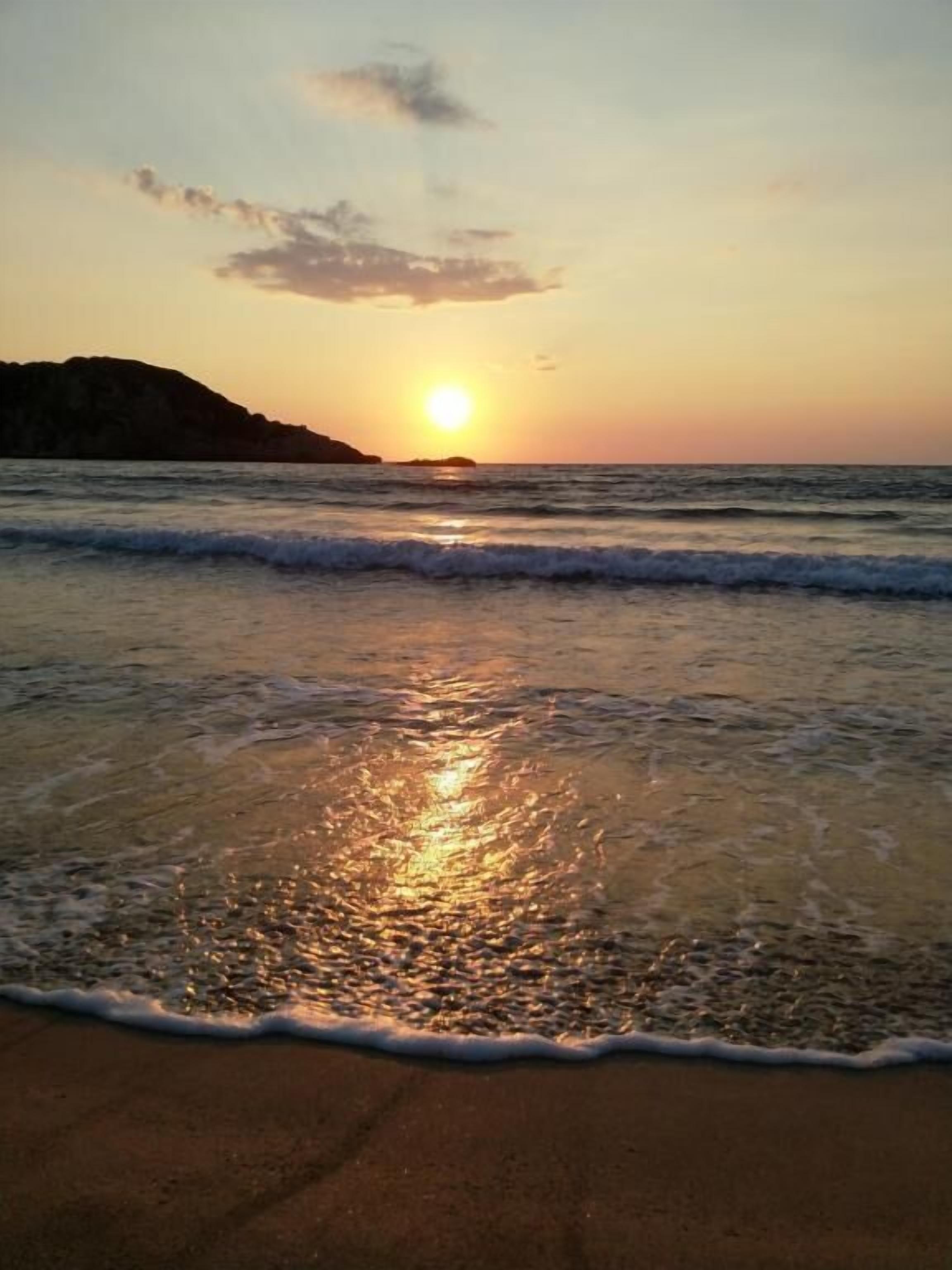 A beach scene with the sun setting over the ocean, waves gently lapping at the shore.