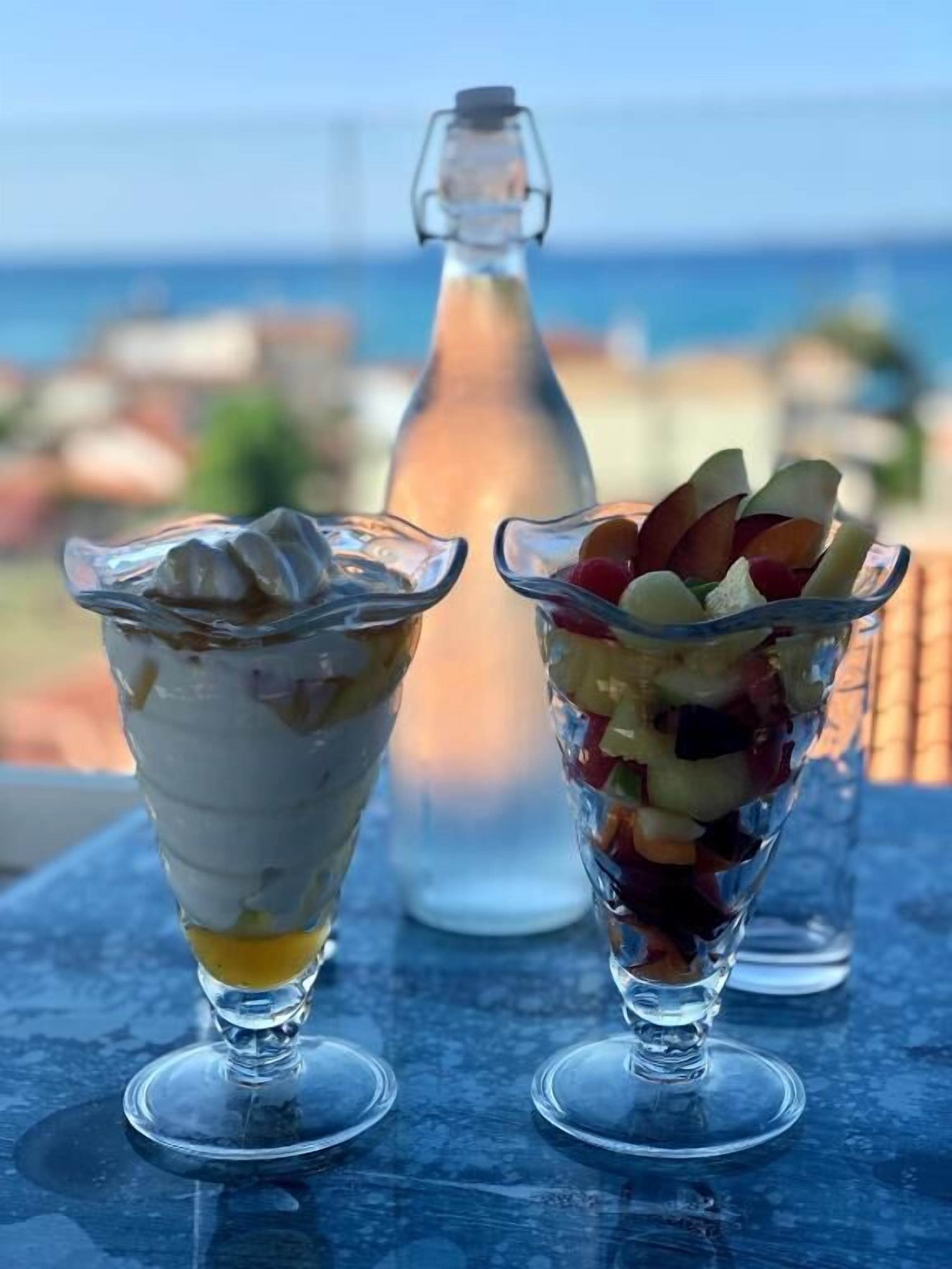 Dos vasos de postre sobre una mesa, con una botella y una vista al mar de fondo.