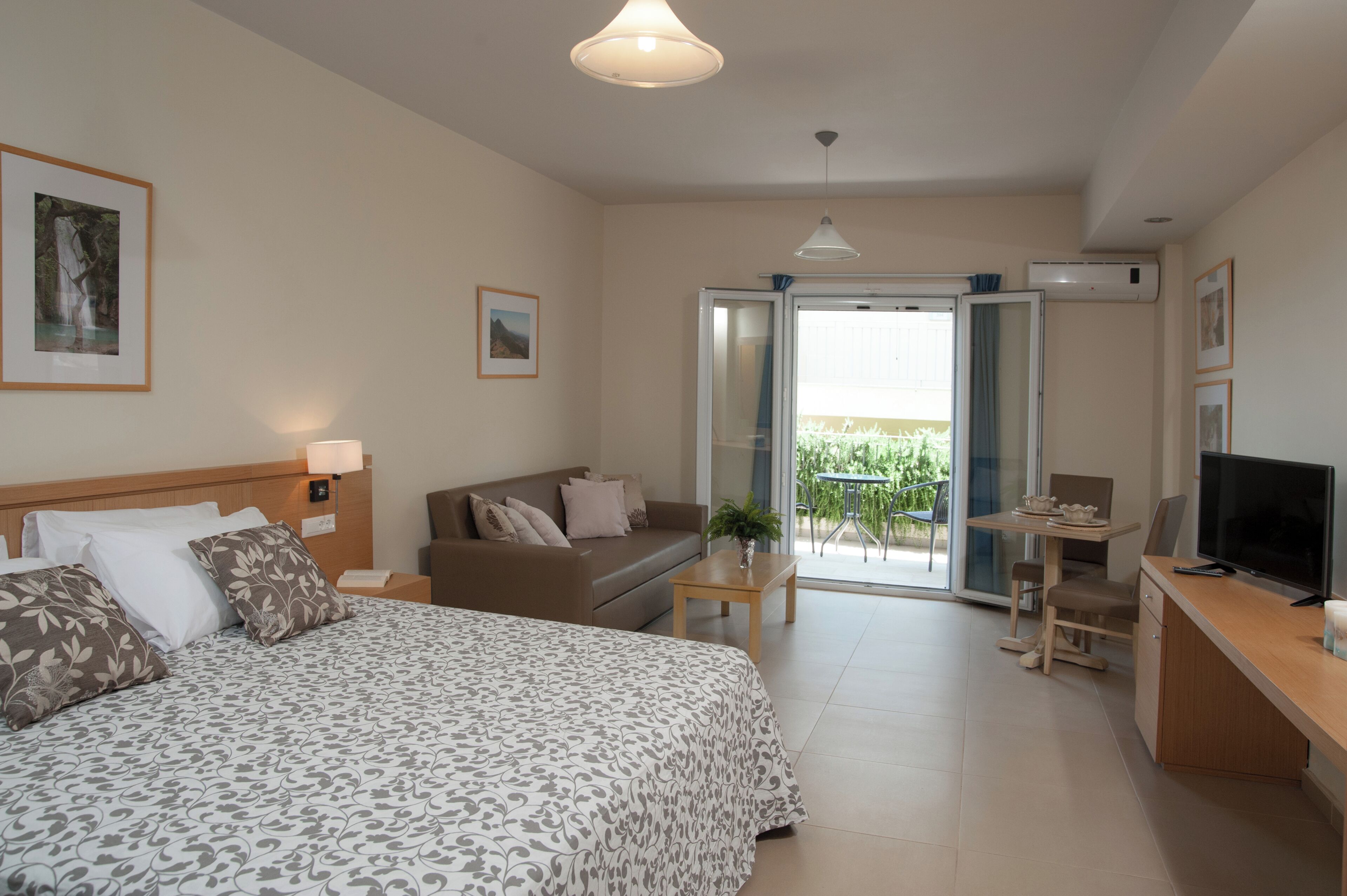 Habitación de hotel con cama, sofá, escritorio con televisor, balcón con mesa y sillas, y unidad de aire acondicionado.