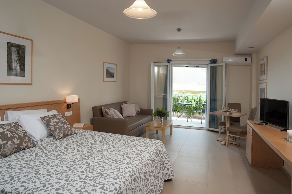 A hotel room with a bed, a sofa, a desk with a TV, a balcony with a table and chairs, and an air conditioning unit.