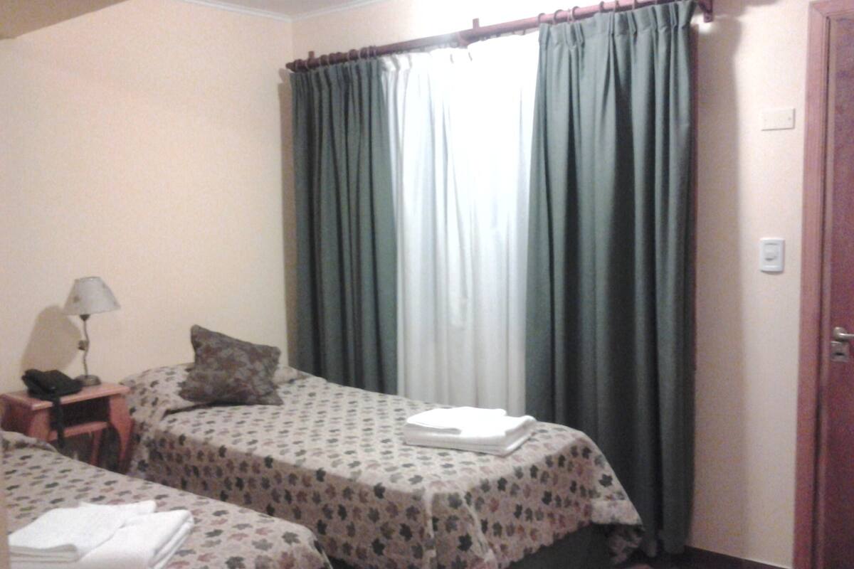 A hotel room with two beds, a nightstand with a lamp, a window with curtains, and a door.