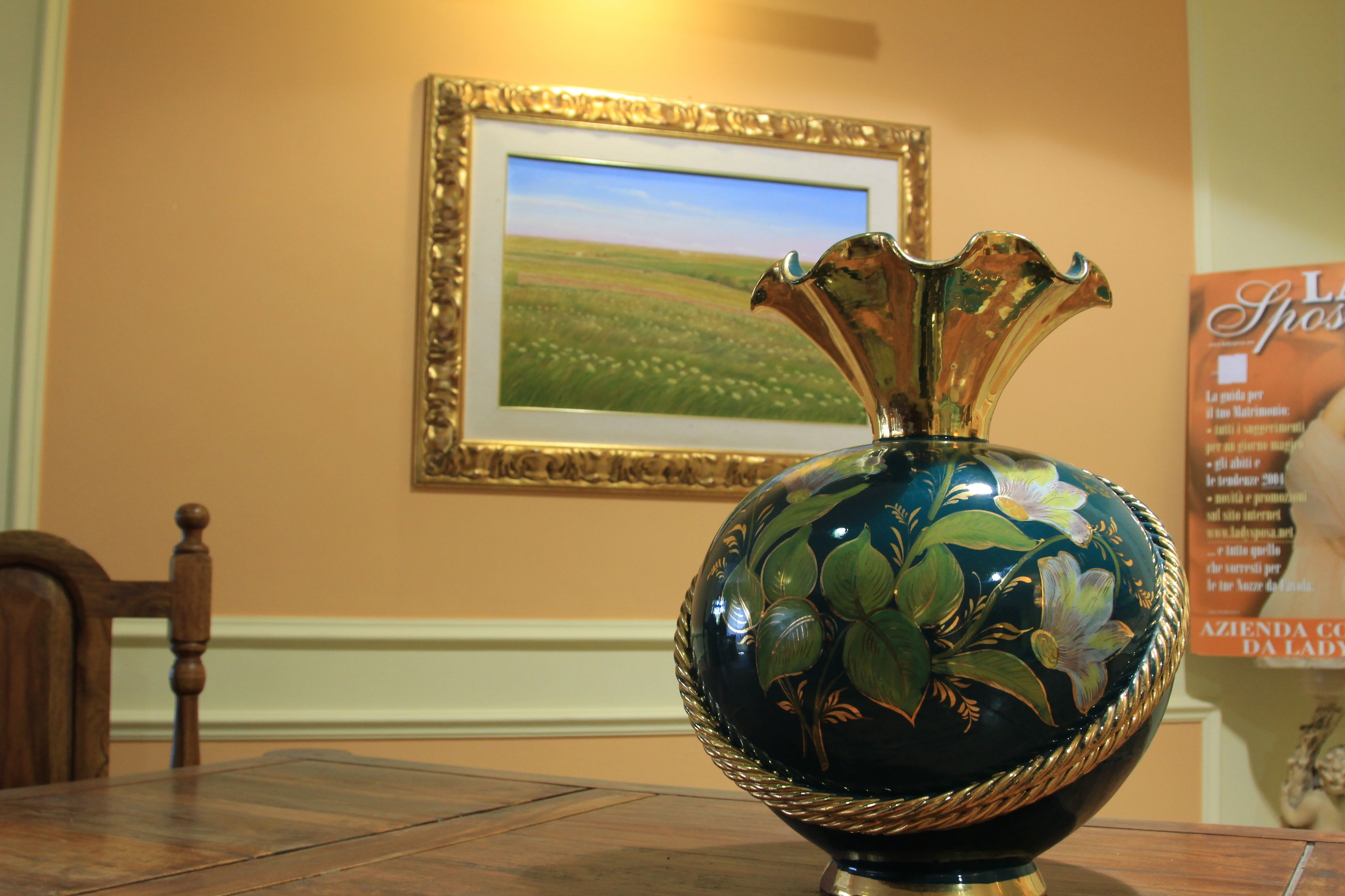 A decorative vase with floral designs on a table in a room with a framed landscape painting on the wall.