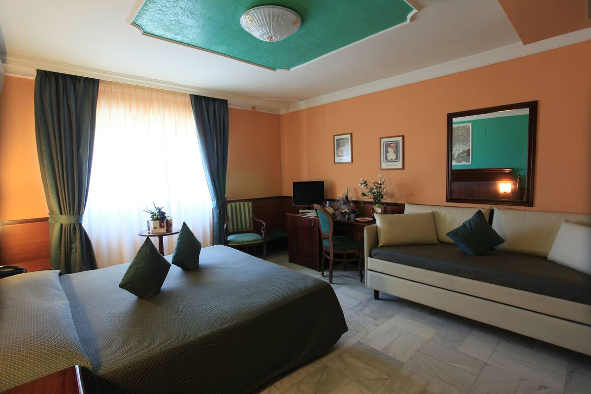 A hotel room with a bed, a sofa, a desk, and a chair. There are curtains, a plant, and a mirror on the wall.