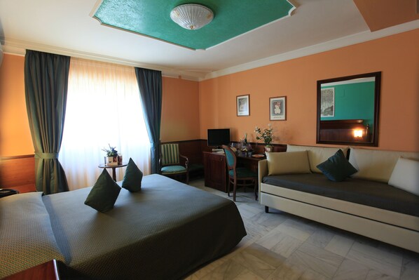 A hotel room with a bed, a sofa, a desk, and a chair. There are curtains, a plant, and a mirror on the wall.