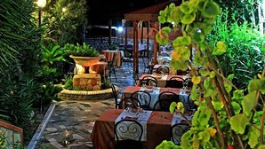 Un área de comedor al aire libre con piso de piedra, mesas con sillas de metal y plantas en macetas.