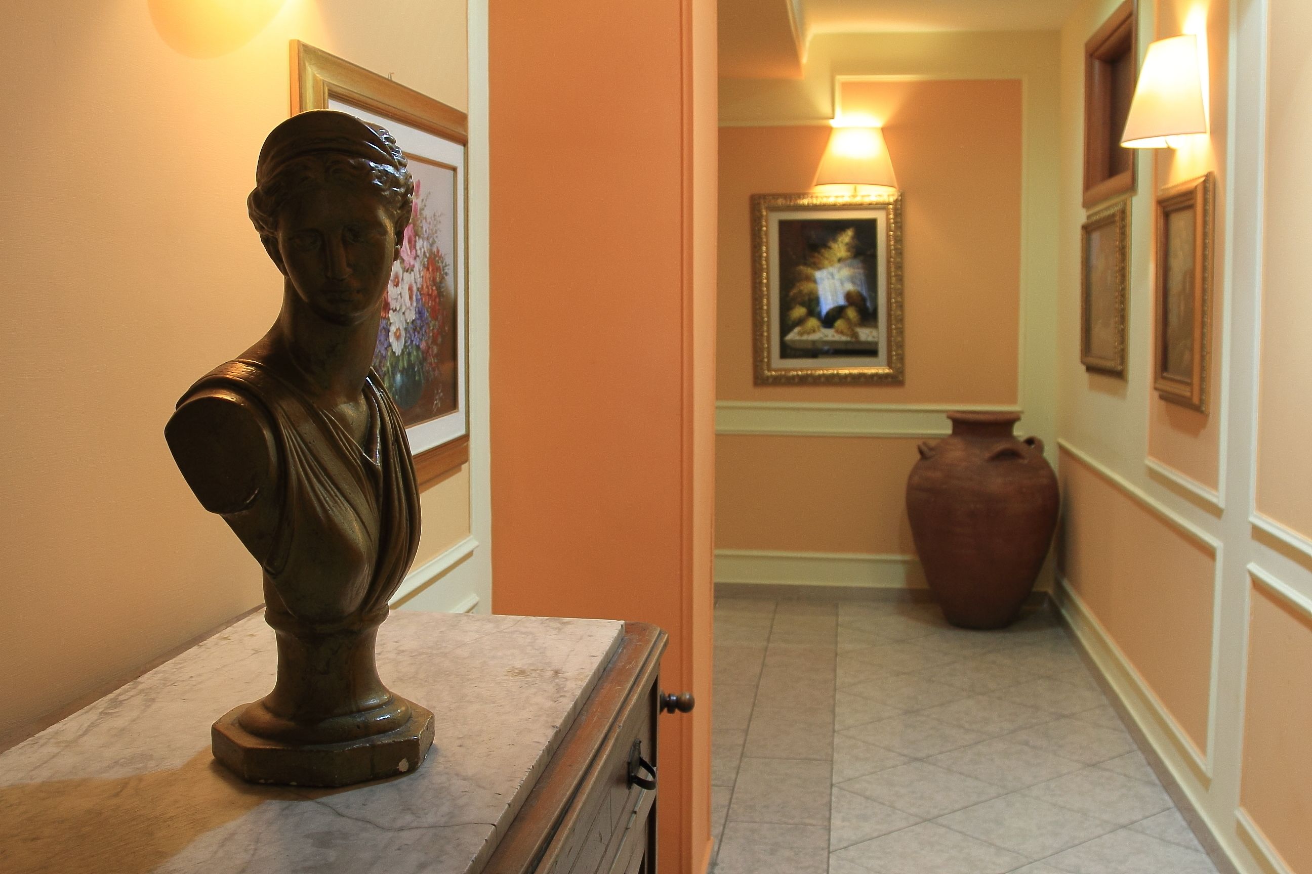 Un pasillo con una escultura de busto sobre un pedestal de mármol, obras de arte enmarcadas en las paredes y un jarrón grande al fondo.