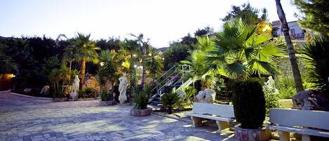 A paved courtyard with stone benches, palm trees, and statues.
