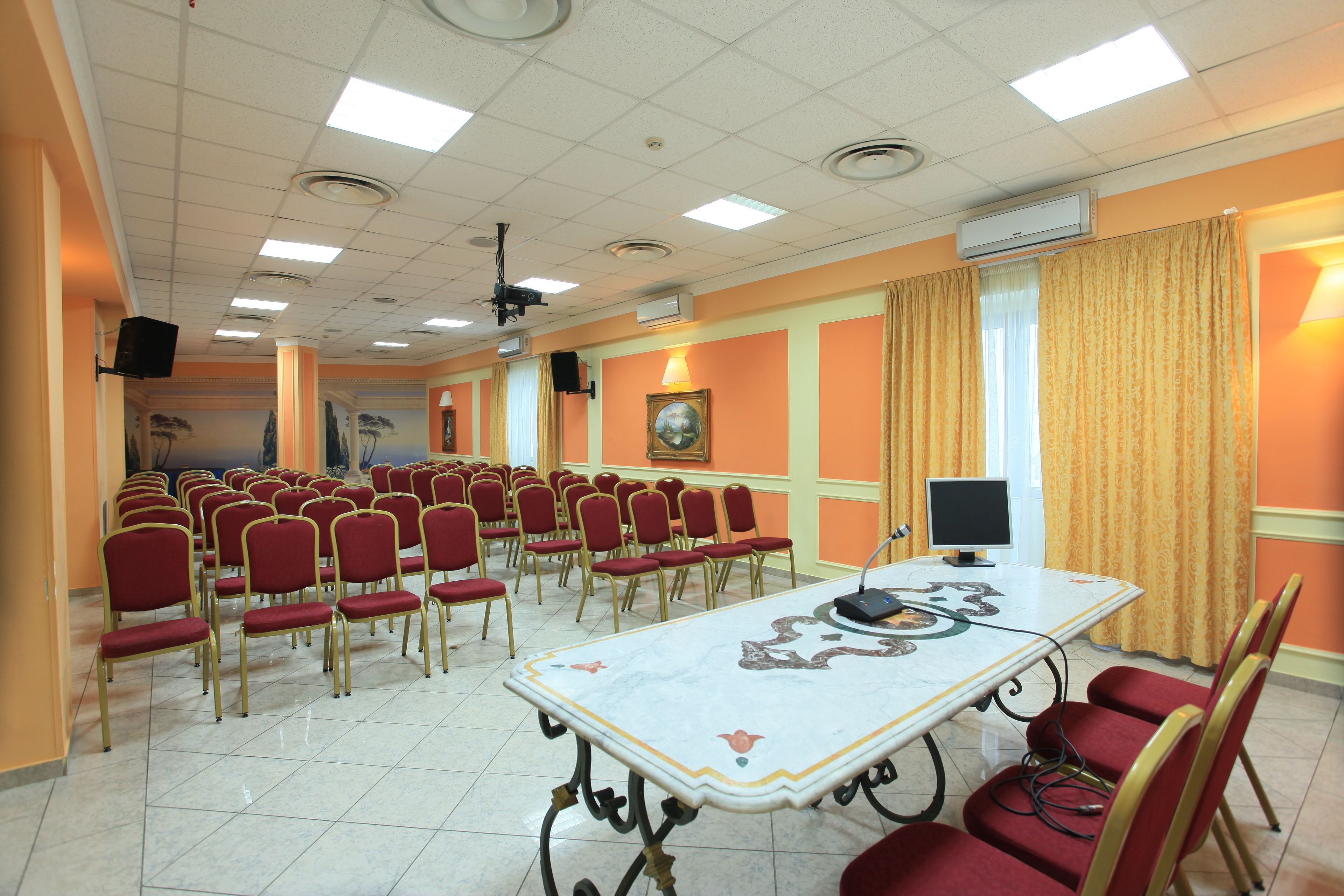 Une salle de conférence avec des rangées de chaises rouges, une grande table recouverte d’une nappe décorative et un écran de projection.