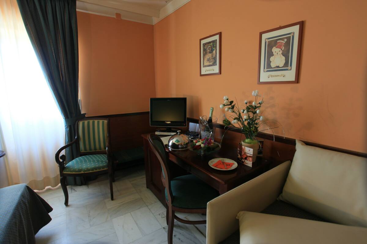 A hotel room with a desk, a television, a chair, a sofa, and two framed pictures on the wall.