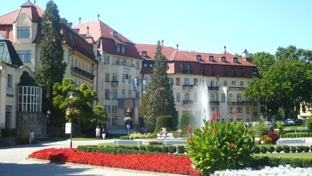 Un grand bâtiment historique à plusieurs étages, surmonté de toits rouges, entouré de jardins bien entretenus et agrémenté d’une fontaine.