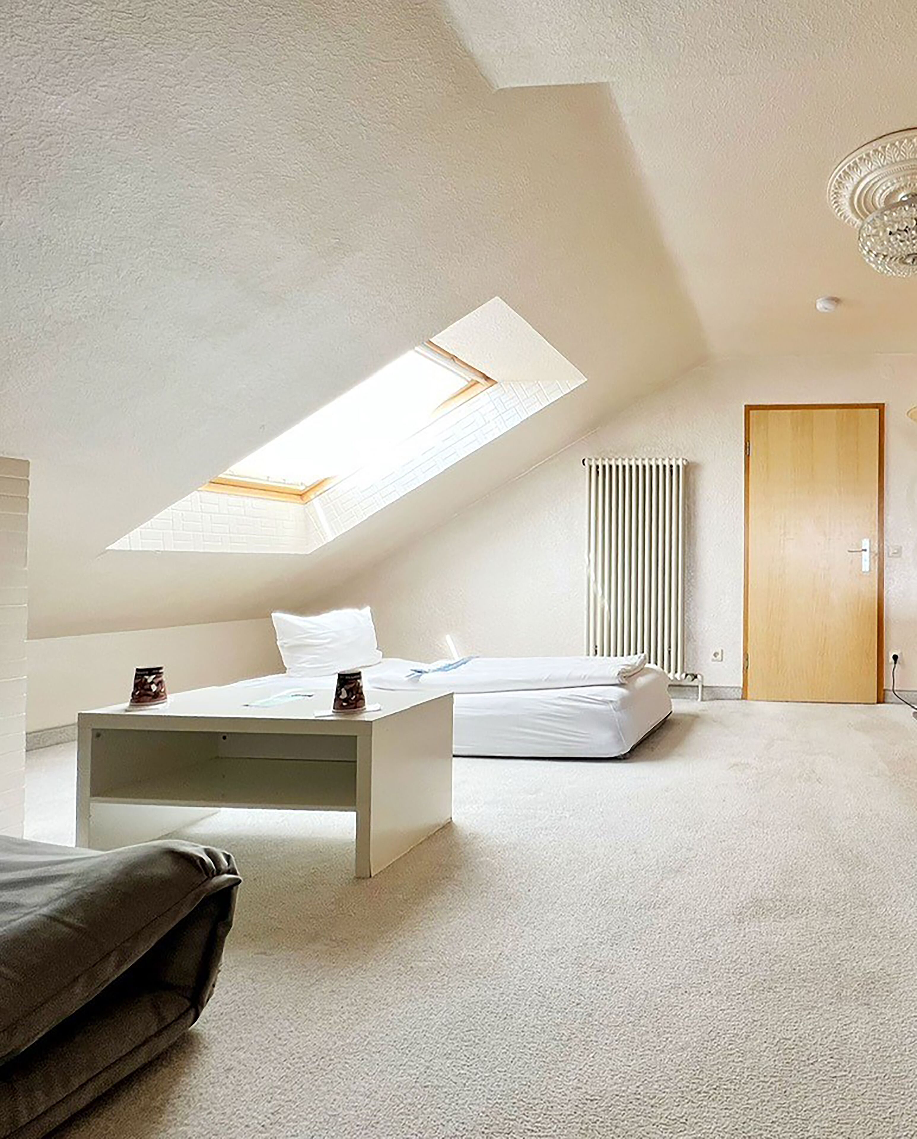 Une chambre à coucher moderne et minimaliste, dotée d’un plafond en pente, d’un puits de lumière, d’un lit, d’une table de chevet et d’un radiateur.