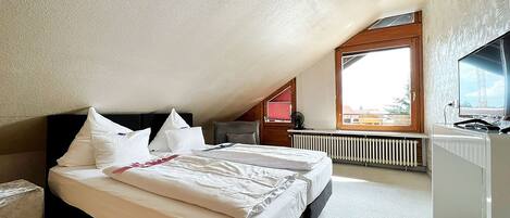 A bedroom with a sloped ceiling, a bed with white linens, a wooden headboard, a radiator, and a window with a view of buildings.