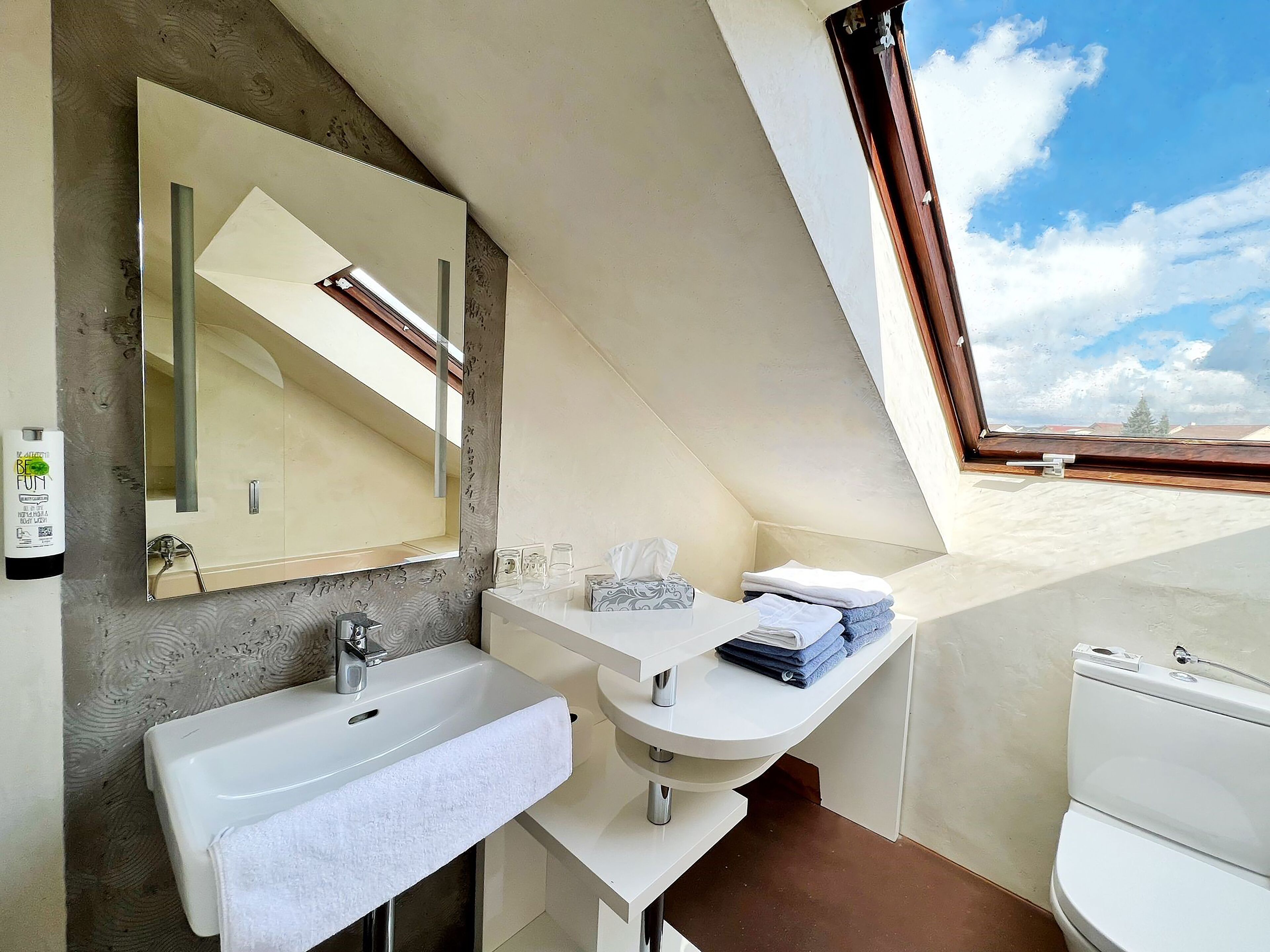 A modern bathroom with a large mirror, a sink, and a toilet. There is a window providing a view of the sky and buildings.
