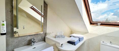 A modern bathroom with a large mirror, a sink, and a toilet. There is a window providing a view of the sky and buildings.