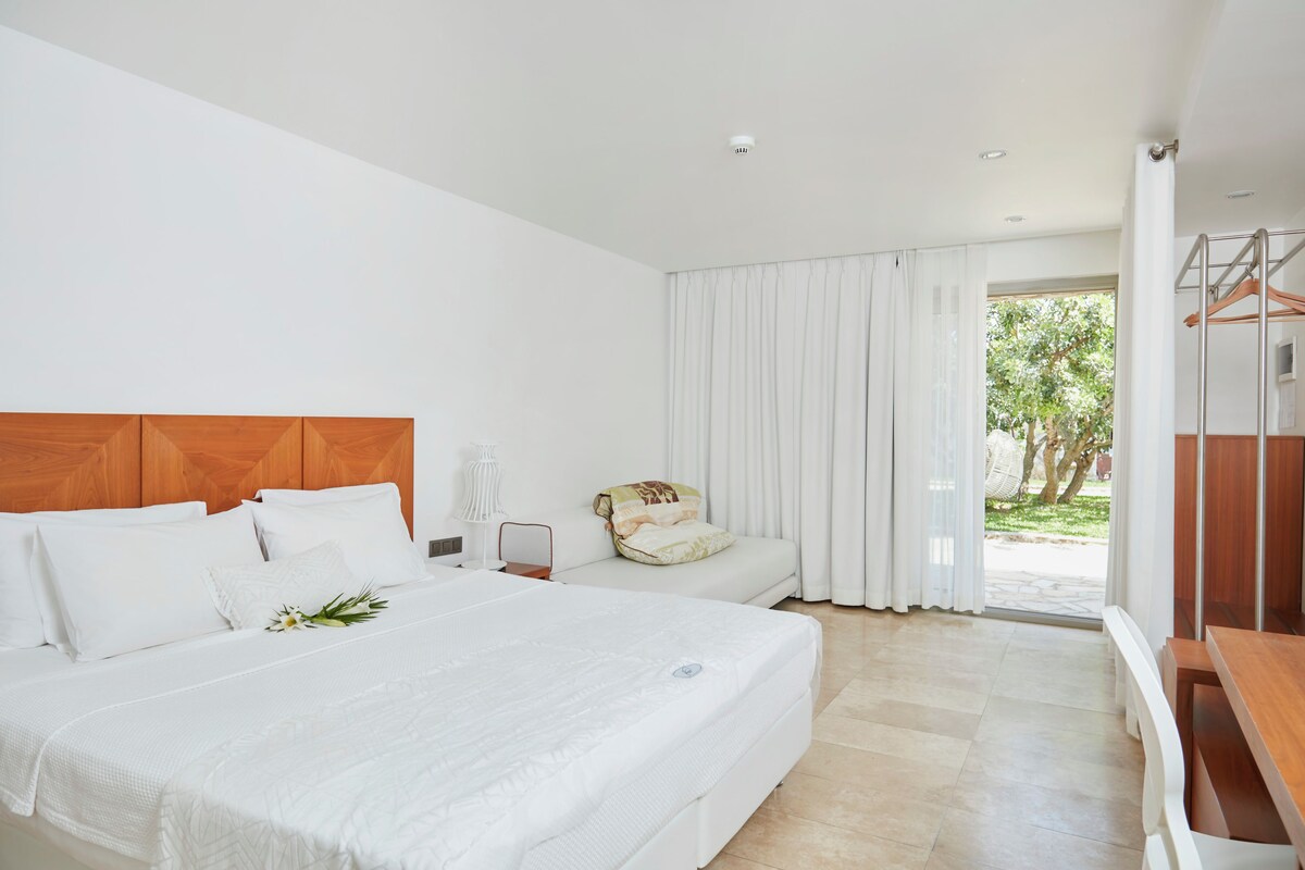 A modern hotel room with a large bed, a wooden headboard, a sofa, and a view of the outdoors.