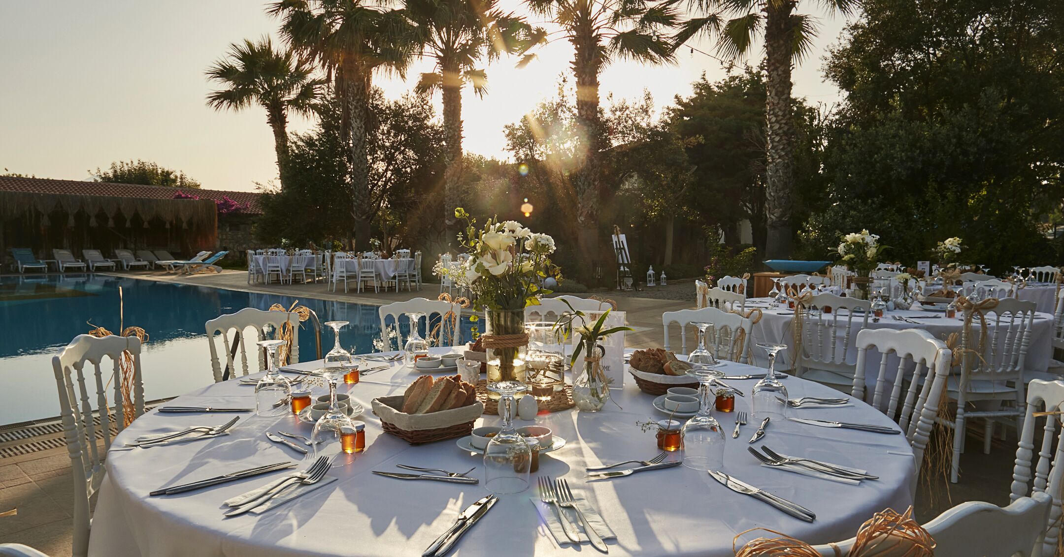 An outdoor dining area with round tables set for a meal, surrounded by chairs, with a pool and palm trees in the background.