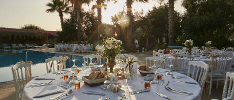 An outdoor dining area with round tables set for a meal, surrounded by chairs, with a pool and palm trees in the background.