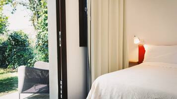 Et hotelværelse med en seng, et natbord med lampe, en stol og udsigt til grøn vegetation udenfor.