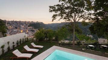 A swimming pool with lounge chairs, a view of a cityscape, and a tree in the foreground.