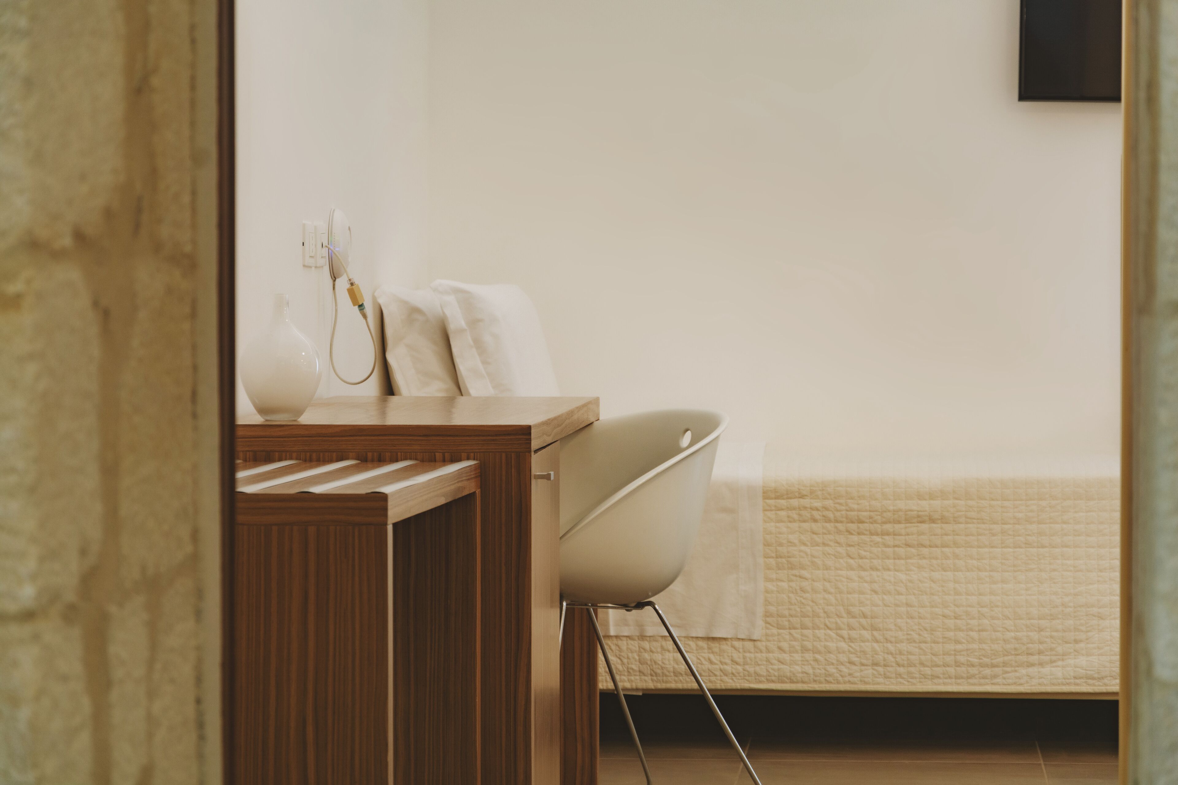 Un dormitorio con un escritorio de madera, una silla blanca, una cama con colcha color beige y un televisor montado en la pared.